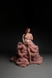 Expecting mother with long red hair wearing an elegant blush pink maternity gown, softly looking down while cradling her baby bump during a fine art studio maternity photoshoot on a dark background by Kaitlyn Dawn Photography.