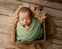 Sleeping newborn baby boy wrapped in a green swaddle and posed in a rustic wooden crate during a studio newborn photography session by Yucaipa newborn photographer Kaitlyn Dawn Photography.
