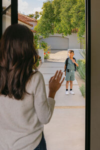 Parent waving goodbye from the doorway as her son leaves for school