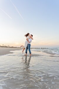 Professional photography by Nature's Child Photography: In Palm Coast, an engaged couple laughs as he spins her barefoot in the water during a beach engagement, their reflection rippling beneath them.