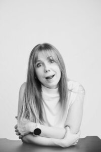 A black and white picture of a blonde woman with straight hair speaking in front of a white backdrop. She wears a white white shirt and an amused expression. Her arms rest on the black table in front of her.