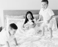 Black and white candid portrait of two energetic brothers jumping on the bed while mom snuggles their newborn baby during an in-home session.