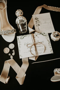 Elegant wedding details showcased against a black background, featuring the bride's perfume, shoes, wedding rings, and invitations.