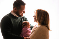 A backlit picture of two parents holding their newborn daughter. The daughter is wrapped in purpled fabric and cradled between her parents. The mom and dad smile at eachother.
