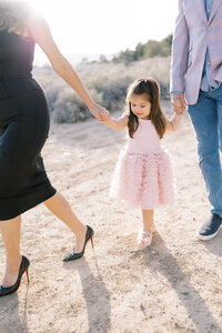The Parker family walking together in the Las Vegas desert, with their young daughter holding both parents’ hands, captured in a soft, sunlit candid family portrait session.
