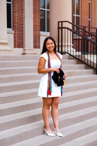 Master's degree senior wearing white dress and heels standing outside on staircase with her cap and cords