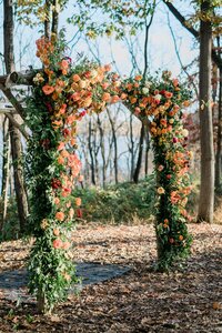 A wooden wedding arch in a forest setting, adorned with vibrant orange, yellow, and red flowers with greenery