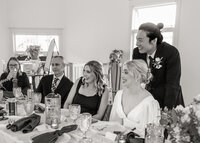 Black and white candid, documentary style photo of a couple and their friends sitting at the reception table at their wedding laughing and having fun with their guests at Mildred Hall in Portland Oregon.