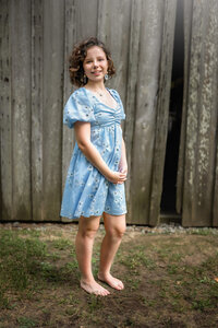 Young girl in light blue dress smiling in front of rustic wooden barn during family photo session in Eastern North Carolina