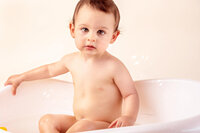 calm toddler sitting in white bathtub with soft bubbles and neutral backdrop