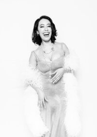 Fine art portrait from Luxe Portraiture LV showcasing a glamorous black‑and‑white image of a woman in a sleeveless metallic mesh dress with feathered shawl, elegant jewelry, and radiant smile, evoking vintage Hollywood style in luxury portrait photography.