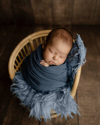 Sleeping newborn baby boy wrapped in a navy swaddle and posed in a wicker chair with soft blue textures during a studio newborn photography session by Yucaipa newborn photographer Kaitlyn Dawn Photography.