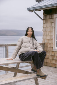 Model in a taupe down puffer and charcoal corduroy trousers seated on a picnic table by the boathouse