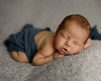Peacefully sleeping newborn baby boy posed on a textured gray blanket with hands tucked under chin during a professional newborn photography session by Yucaipa newborn photographer Kaitlyn Dawn Photography.