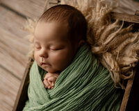 Sleeping newborn baby boy wrapped in a sage green swaddle and resting in a wooden crate with soft neutral textures during a newborn photography session by Yucaipa newborn photographer Kaitlyn Dawn Photography.