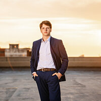 Senior guy posing in a blue suit with the Mobile, Alabama sunset shining behind him.