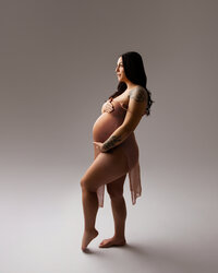 Pregnant woman standing in profile wearing a fitted neutral maternity gown during a minimalist studio pregnancy photography session captured by Kaitlyn Dawn Photography.
