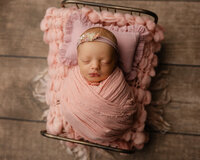 Sleeping newborn baby girl wrapped in a blush pink swaddle and posed on a textured blanket during a studio newborn photography session with newborn photographer Kaitlyn Dawn Photography.