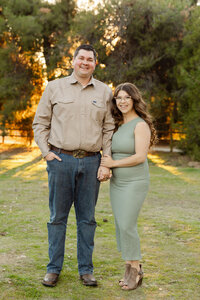 Parents standing together in a grassy park during golden hour, coordinated neutral outfits and warm sunset light captured by Kaitlyn Dawn Photography, Yucaipa family photographer.