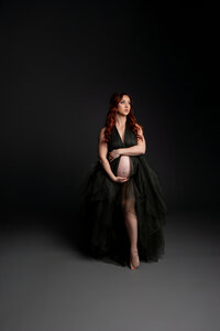 Pregnant woman with long red hair wearing a flowing dark green tulle maternity gown while standing barefoot and gently holding her baby bump during a dramatic studio maternity photoshoot on a dark backdrop captured by Kaitlyn Dawn Photography.