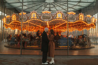 A bride and groom at their engagement session in DUMBO Brooklyn, standing in front of the carousel, holding hands in Brooklyn Bridge Park. Parr Photo Co.
