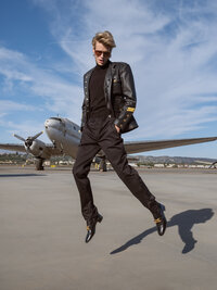 Person in a black leather jacket and sunglasses caught mid-jump on an airfield tarmac, propeller aircraft behind.