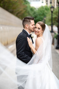 Bride and groom embrace while wedding veil flows in the wind