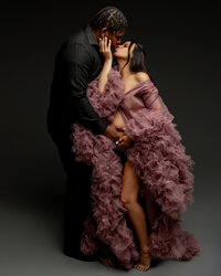 Expecting couple sharing a kiss during a dramatic studio pregnancy photography session with the mother wearing a flowing mauve ruffle gown photographed by Kaitlyn Dawn Photography.