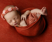 Newborn baby wrapped in deep red fabric wearing a matching floral headband. Photographed by Inland Empire newborn photographer Kaitlyn Dawn Photography.
