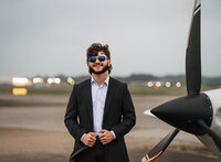 Senior portrait at a small hangar near Mobile International Airport in Mobile, Alabama — suit
jacket over his shoulder, wearing aviators with the plane in the background.