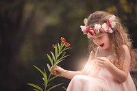 Young girl wearing a floral crown and soft pink dress looks down in surprise as a tiny glowing fairy dances near her lap during a fairy adventure signature in Palatka. (Photographed by Nature's Child Photography)
