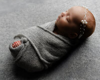 Sleeping newborn baby girl wrapped in a textured gray swaddle and wearing a pearl headband while posed on a soft neutral backdrop during a classic newborn photography session by Kaitlyn Dawn Photography.