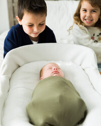 Older brother and sister smiling and watching their newborn sibling resting in a bassinet during an in-home newborn session.