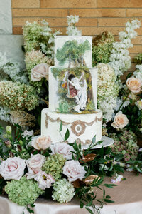 A three-tier luxury wedding cake featuring intricate hand-painted classical art and a base of lush summer roses.