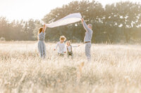 A family photo of mom an dad holding a blanket up in the air as their boys run under in the middle of a DFW field.
