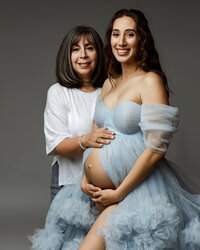 Pregnant woman and older woman posing together in a studio, celebrating pregnancy with a blue gown session by Kaitlyn Dawn Photography in Yucaipa.