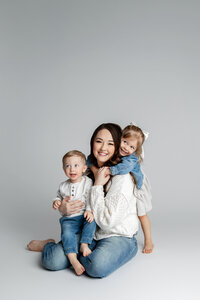 Mother sitting on studio floor holding her two young children close, soft neutral backdrop and natural light creating a warm family portrait by Kaitlyn Dawn Photography, studio family photography specialist.