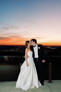 Bride and groom share a kiss on the terrace at Burritt on the Mountain with a vibrant sunset and city lights in the background.