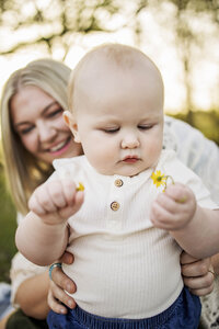 A one year old boy hold a wild flower while his mom looks on and laughs
