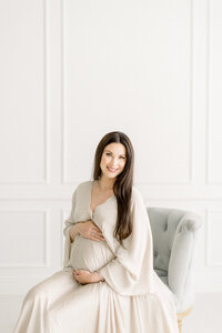 A beautiful portrait of a pregnant Dallas mother sitting on a chair holding her belly as she poses for her maternity session at a Dallas photography studio.