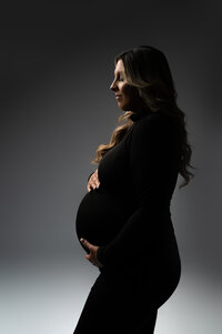 Elegant side profile of a pregnant woman in a black turtleneck, softly illuminated with moody directional lighting during a minimalist maternity session near New Berlin, Wisconsin.