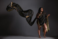 A woman draped in a flowing black and gold sari stands barefoot against a dark background. Her arm is raised, making the fabric billow dramatically—an image perfect for fashion-inspired editorial portraits Washington DC.