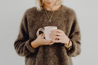 kaboompics_A woman in a brown sweater holds a pink, minimalist mug