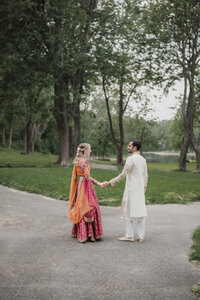 Couple se tenant la main sur un sentier, mariée en lehenga orange et rose éclatant, marié en kurta blanc brodé, entourés de verdure lors de leur cérémonie du Mehndi