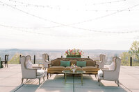 Outdoor lounge seating at Burritt on the Mountain during Callie and Hayes’ wedding, featuring a vintage brown sofa, ivory armchairs, floral arrangements, and string lights with panoramic Huntsville views.