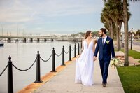 St. Augustine wedding capturing newlyweds strolling along the bayfront, palm trees lining the path and the Bridge of Lions rising in the distance. - A portrait experience by Nature's Child Photography.