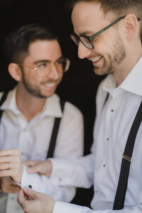Mariés riant ensemble pendant les préparatifs, chemises blanches et bretelles, lunettes et alliance visibles, moment de complicité avant la cérémonie