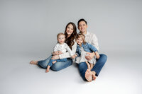 Family of four sitting together in a minimalist studio with light gray background, coordinated neutral outfits and joyful expressions captured by Kaitlyn Dawn Photography, professional family portrait photographer.