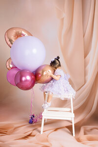 A young girl sits on a white stool hugging her birthday balloons.