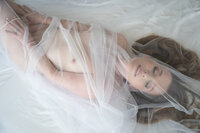 A nude woman lies on a white sheet, delicately covered by a white translucent veil, adding an ethereal quality to the scene